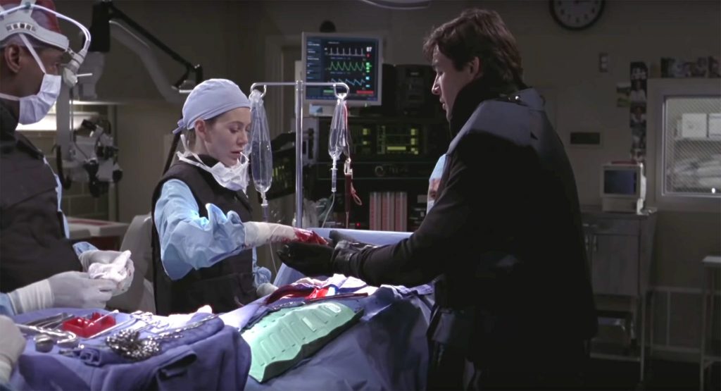 Meredith, Burke and Dylan are in the OR stood over a patient. Meredith has removed the bomb from the patient's chest and is passing it to Dylan.