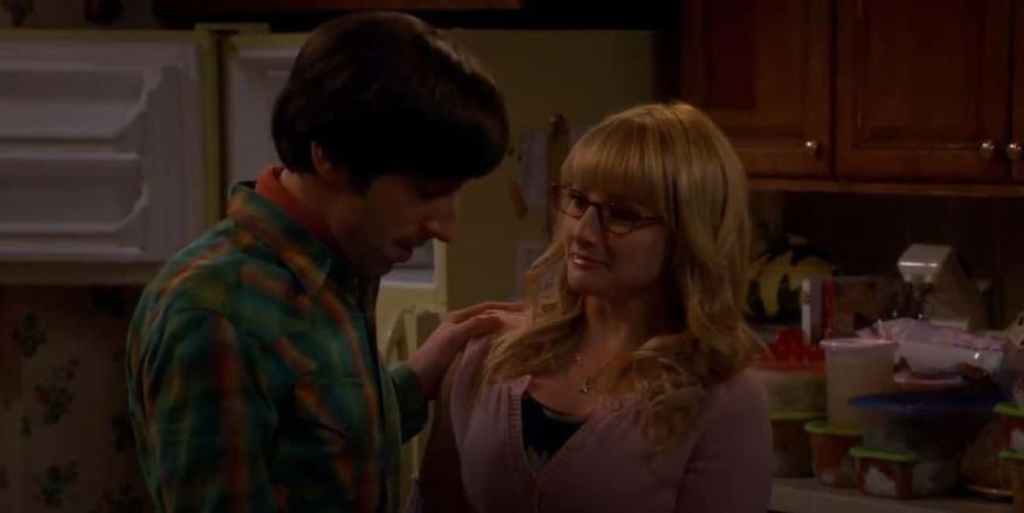 Still from The Big Bang Theory. Stood in the kitchen, an emotional looking Bernadette looks lovingly at Howard who has his head bowed.